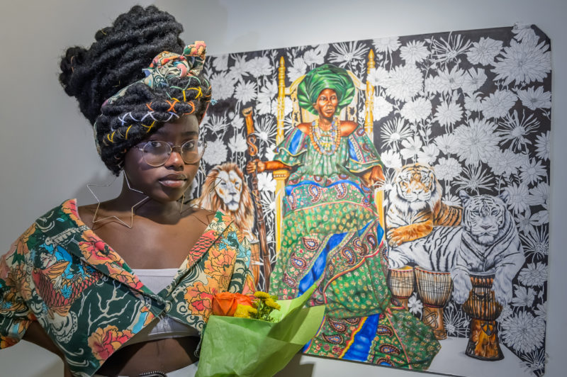 Aisha Mpiana standing in front of artwork featuring seated woman and tigers