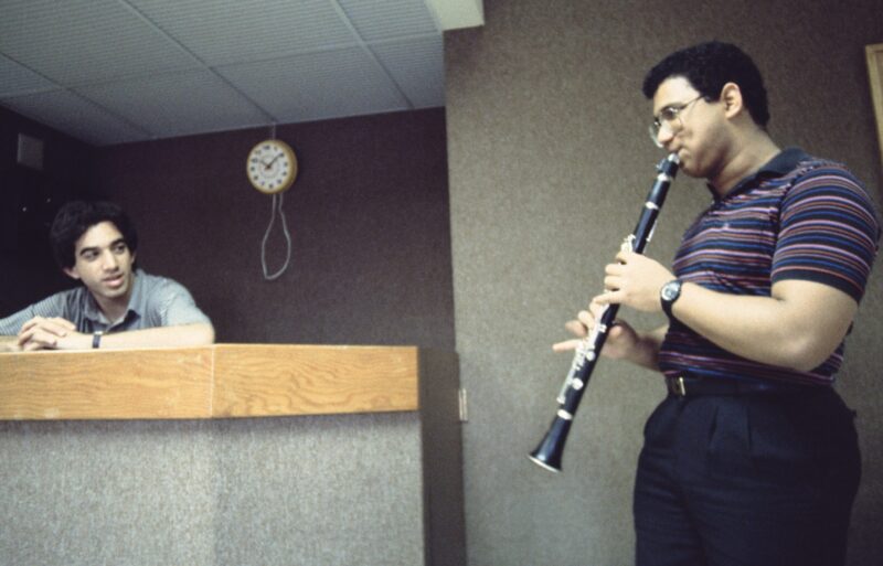 two people, one standing and playing a clarinet, the other watching from behind a counter.