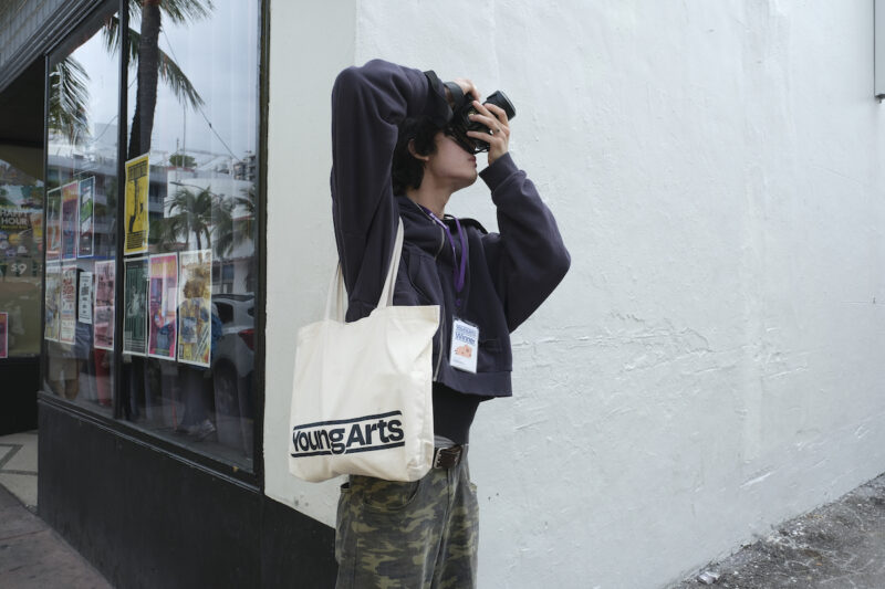 Person with a YoungArts tote bag holding a camera, taking a photograph